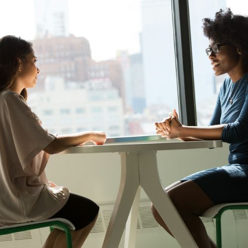 Two women at work, sat at a table facing each other. They are sat next to a window with a city view.