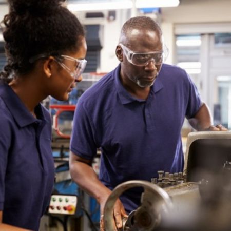 Young woman wearing safety goggles, on an apprenticeship being taught how to use machinery by an older man also wearing safety goggles.